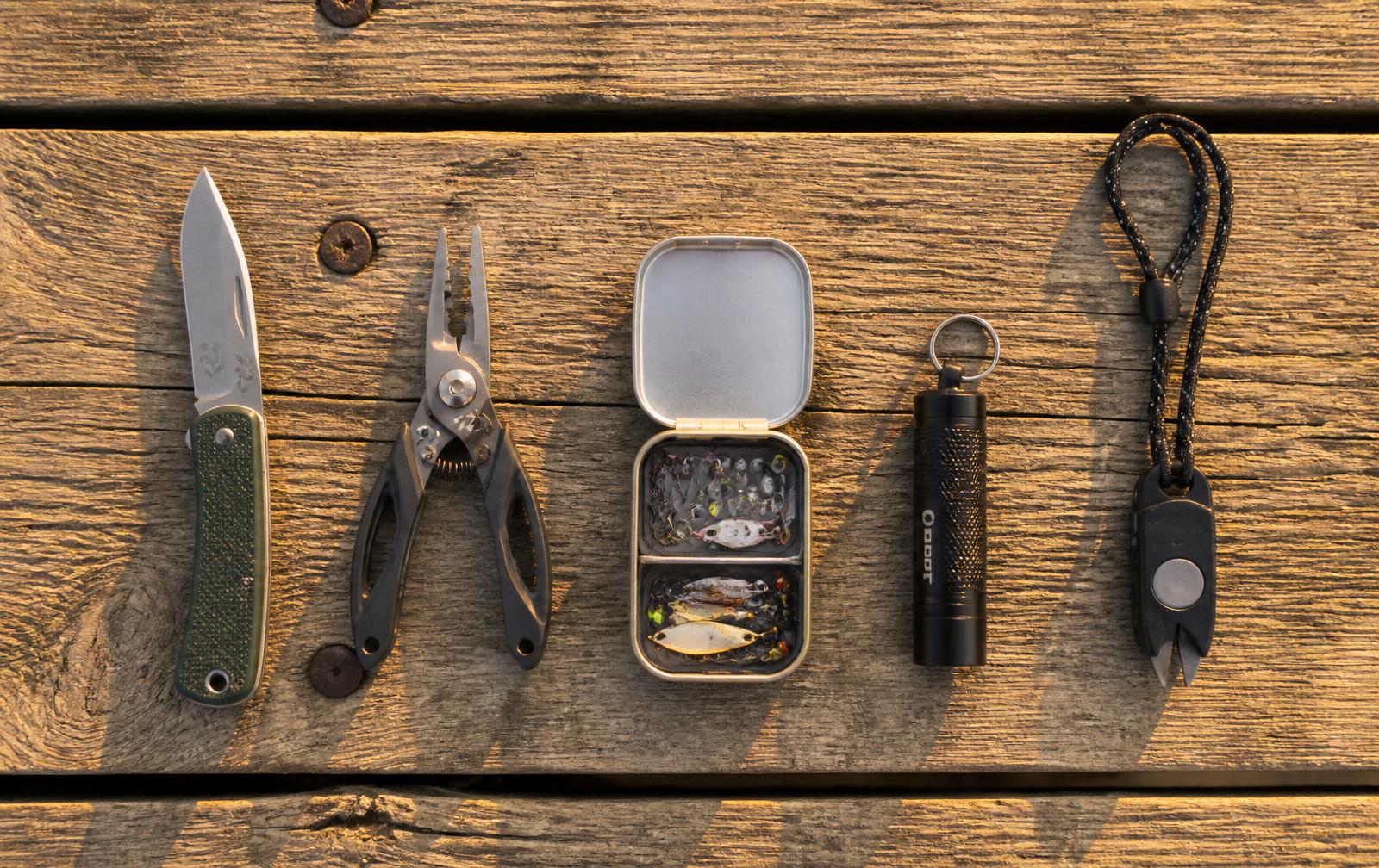Folding fishing knife, mini pliers, tackle tin, keychain light and line clippers on a dock